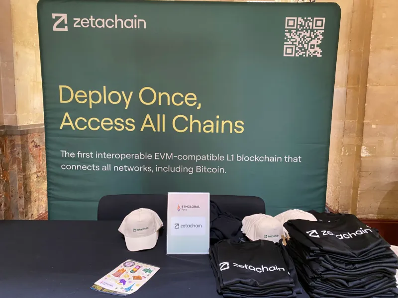 The ZetaChain booth at ETHGlobal Paris, with a banner reading “Deploy Once, Access All Chains” and branded caps and T-shirts on the table